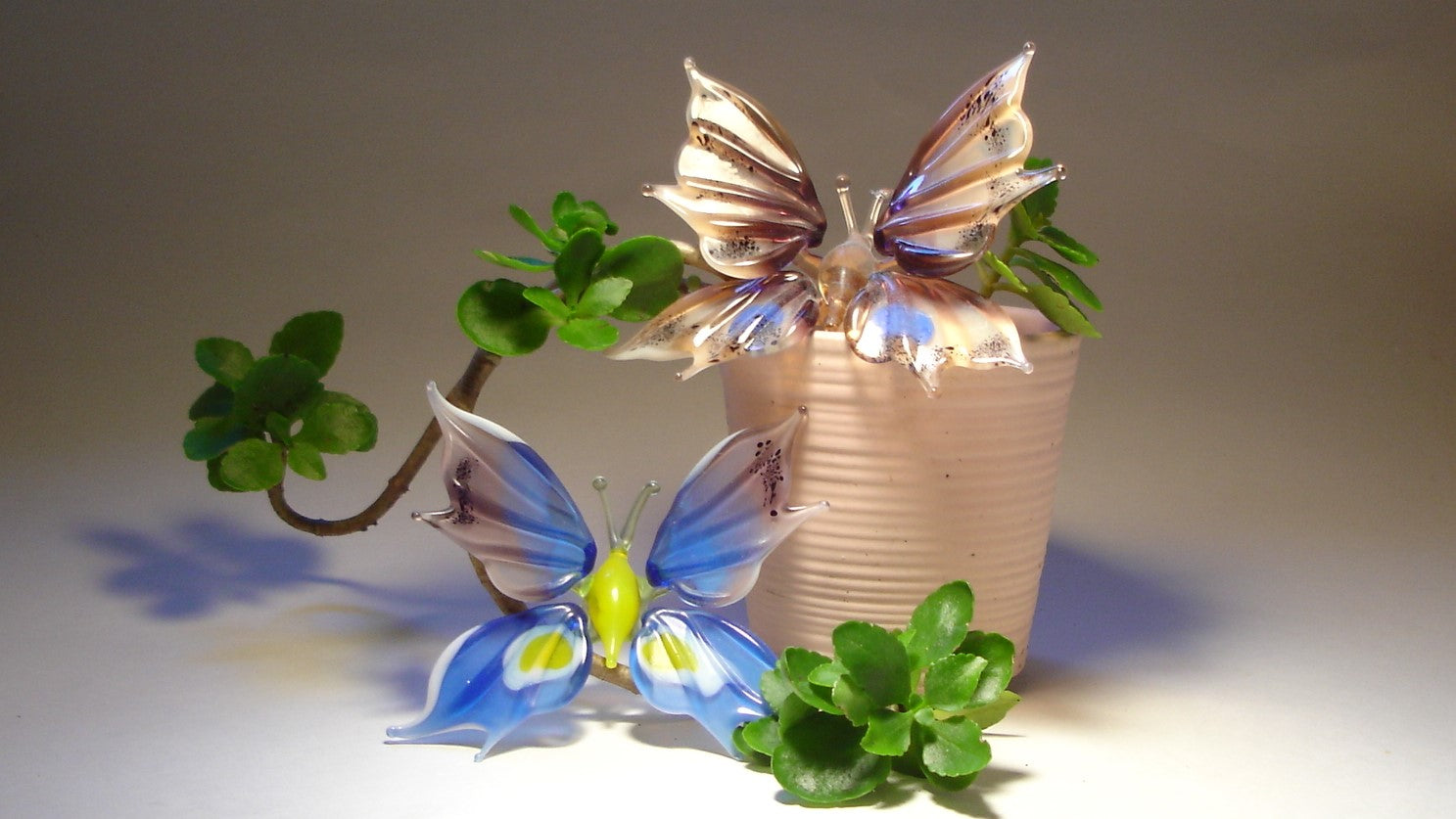 Front view of a beige plant pot and two glass butterflies, one on top, the other one beside the plant.