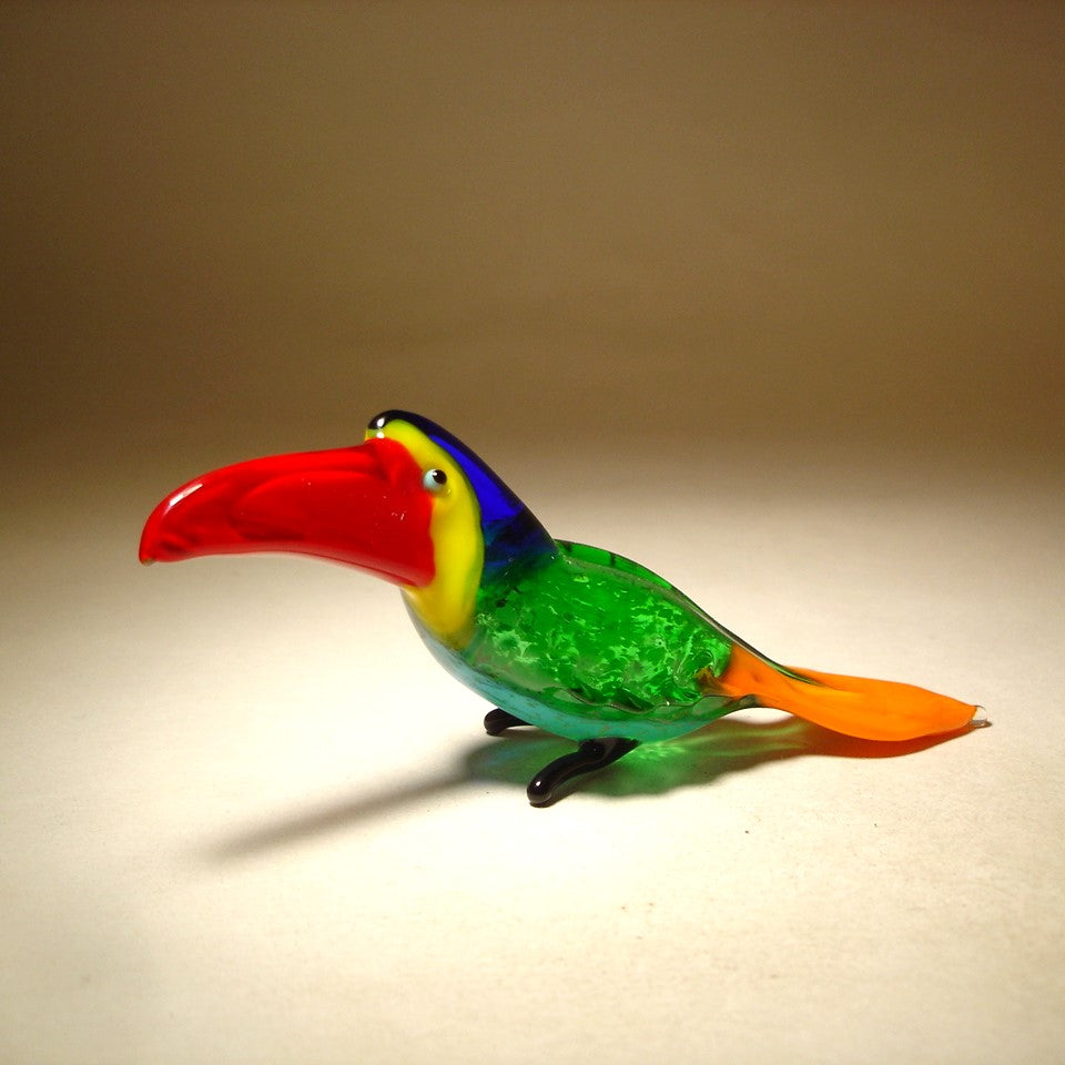 A side view of a glass toucan figurine, emphasizing the bold red beak and the smooth curves of its body.
