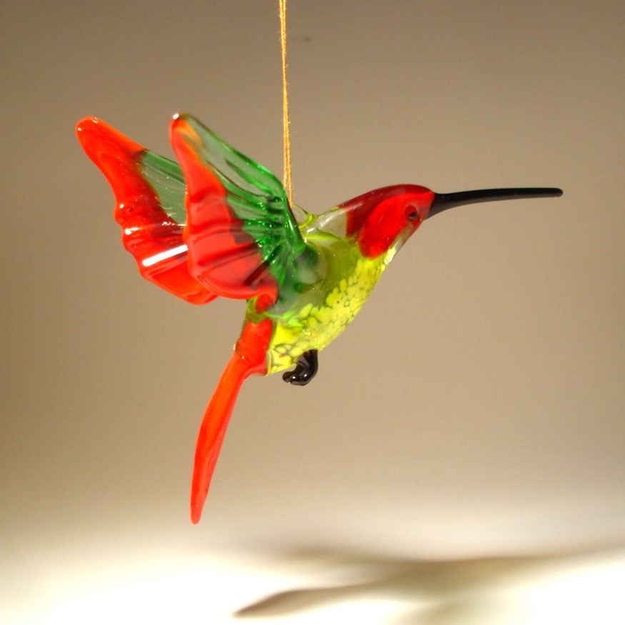 A bright red and yellow glass hummingbird ornament suspended in mid-air, featuring intricate details on its green wings.
