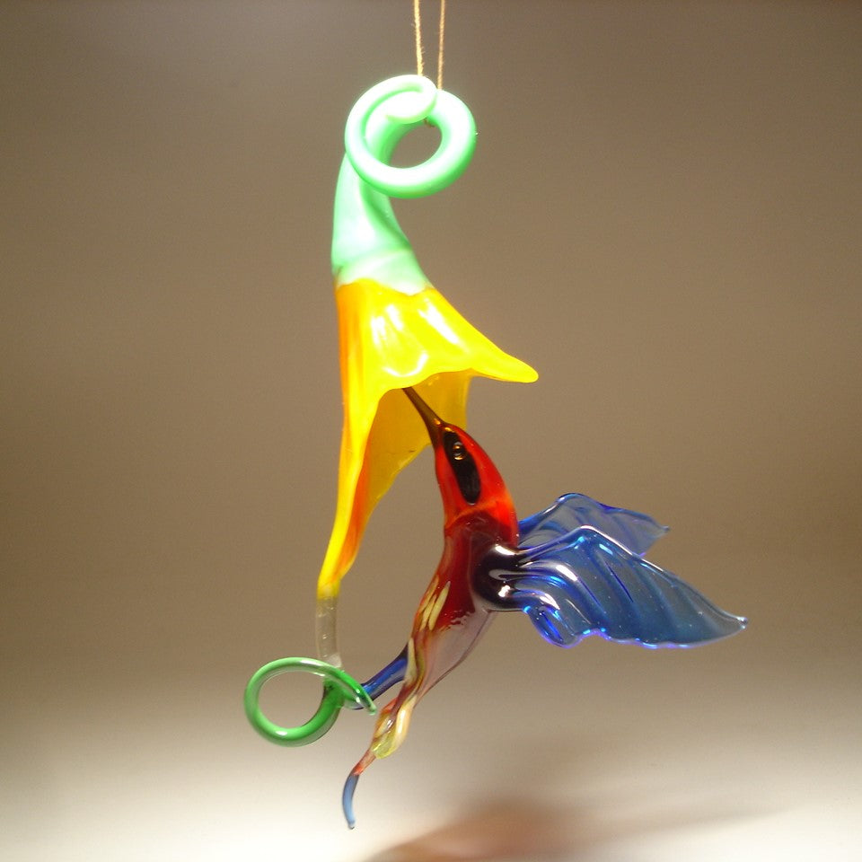 Glass hummingbird ornament hovering near a bright yellow flower, showing delicate details of the bird's wings and floral accents.