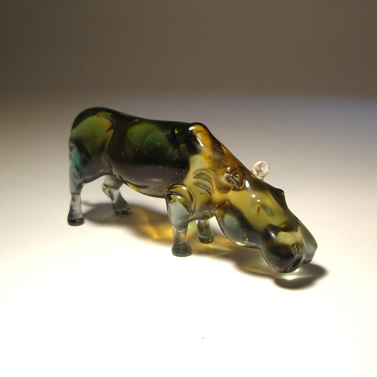 A small hippopotamus figurine made of transparent grey glass standing on a white surface against a neutral gray background.