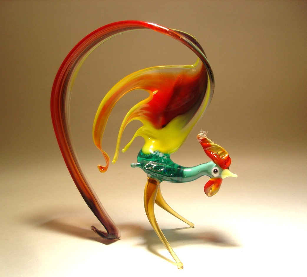 A glass figurine of a rooster with a green body and a glossy black, red and yellow tail that curls elegantly behind it.