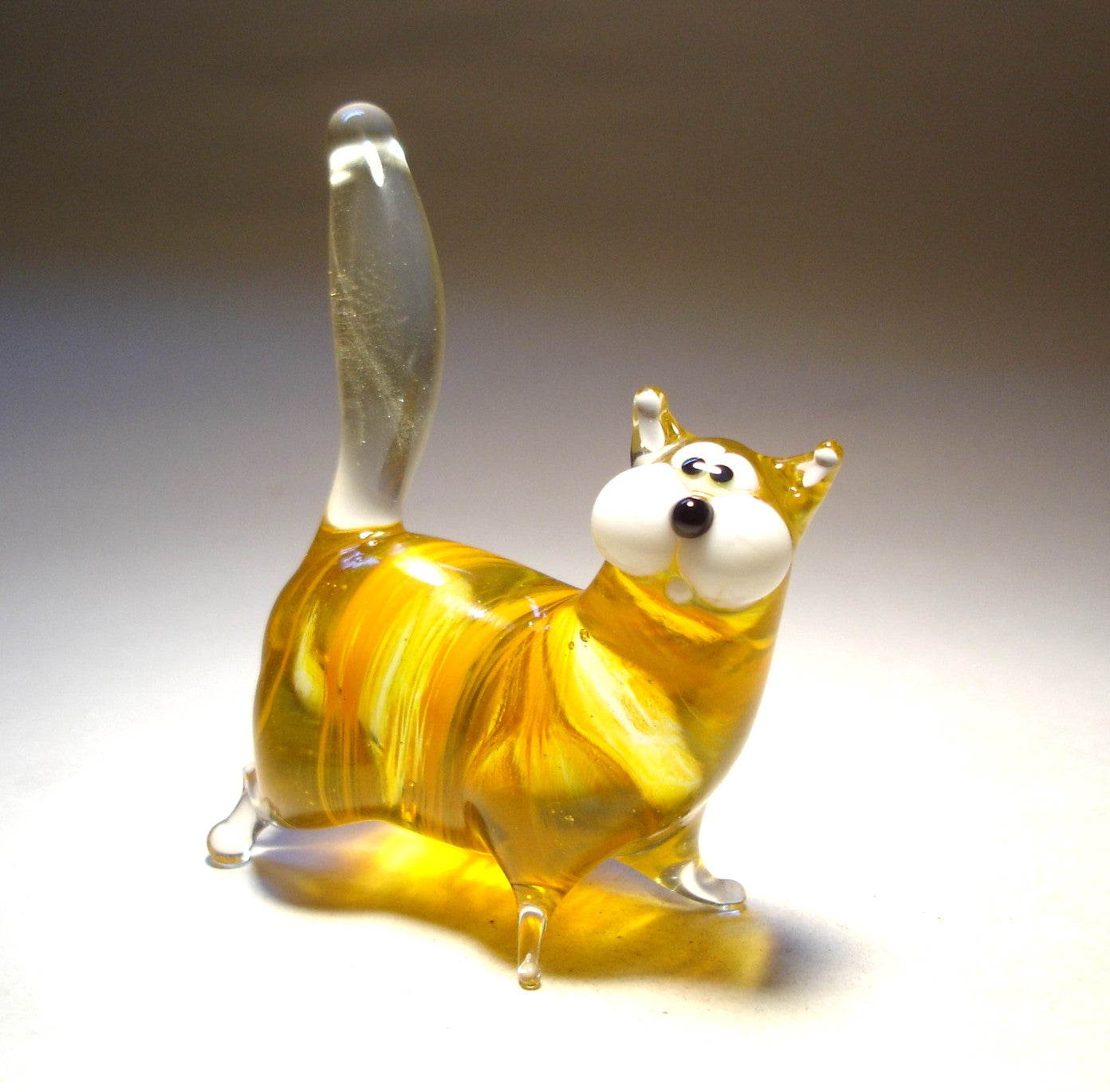 A handmade yellow-orange glass chubby cat figurine, standing with its clear glass tail pointed straight up.
