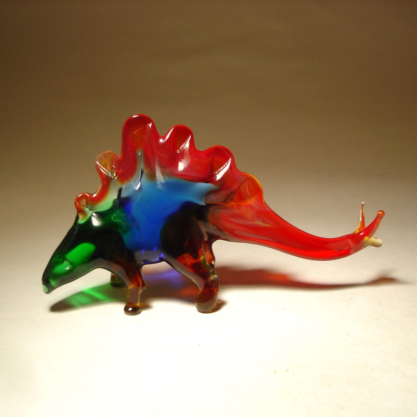 A side profile of the glass Stegosaurus figurine, highlighting its curved tail, arched back, and the row of spiked plates along its spine.