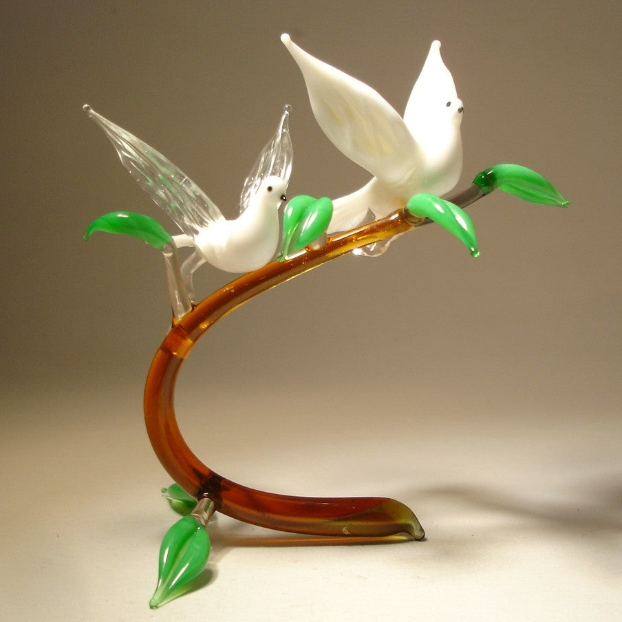 A front view of a glass figurine featuring two white doves perched on a glass branch, showcasing their outstretched wings in an elegant, peaceful pose.