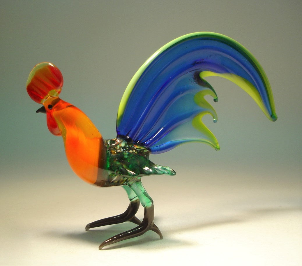 Left side view of a handmade blue glass rooster figurine, showcasing its green body and intricately designed tail feathers