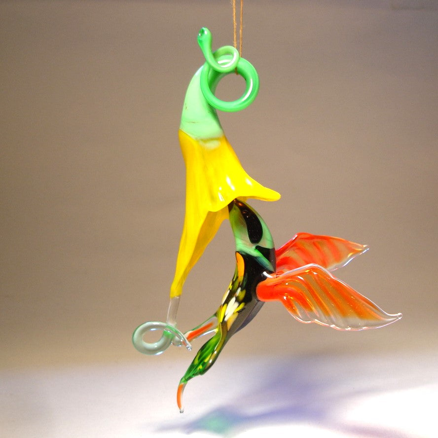 Glass hummingbird ornament hovering near a bright yellow flower, showing delicate details of the bird's wings and floral accents.