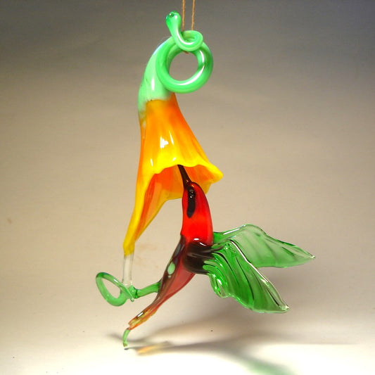 Glass hummingbird ornament hovering near a bright yellow flower, showing delicate details of the bird's wings and floral accents.