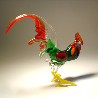 A colorful hand-blown glass rooster figurine with a curved red and clear tail, green body, blue accents, and yellow legs