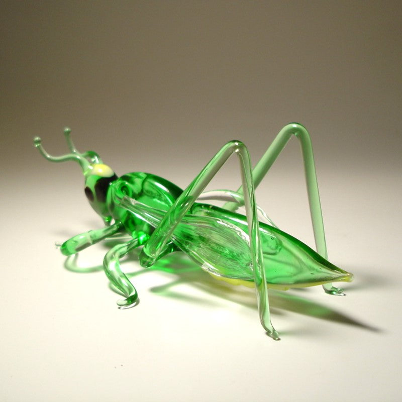 Left side view of a glass cricket grasshopper figurine, displaying its delicate legs, antennae, and light green body.