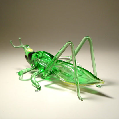 Left side view of a glass cricket grasshopper figurine, displaying its delicate legs, antennae, and light green body.