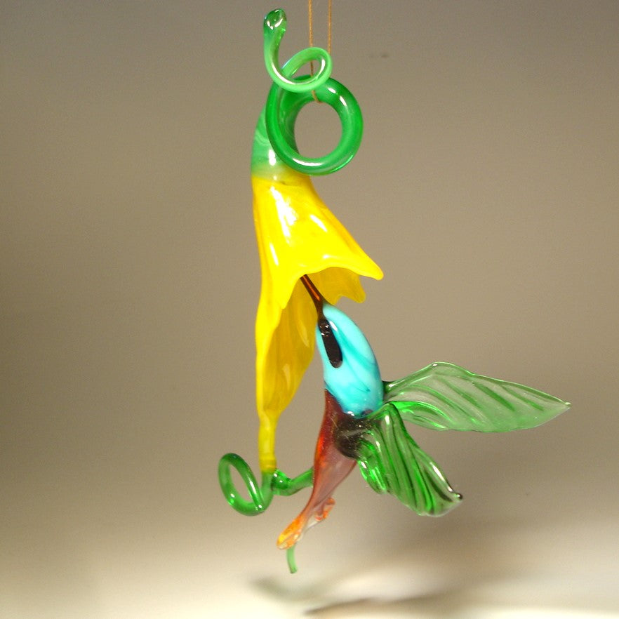 Glass hummingbird ornament hovering near a bright yellow flower, showing delicate details of the bird's wings and floral accents.