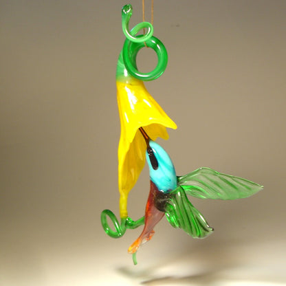 Glass hummingbird ornament hovering near a bright yellow flower, showing delicate details of the bird's wings and floral accents.