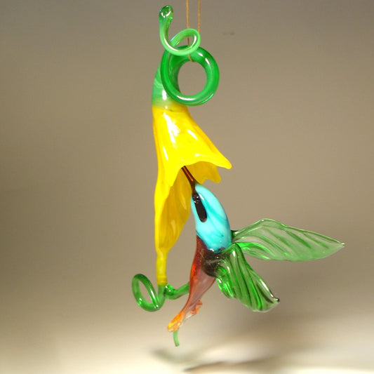 Glass hummingbird ornament hovering near a bright yellow flower, showing delicate details of the bird's wings and floral accents.