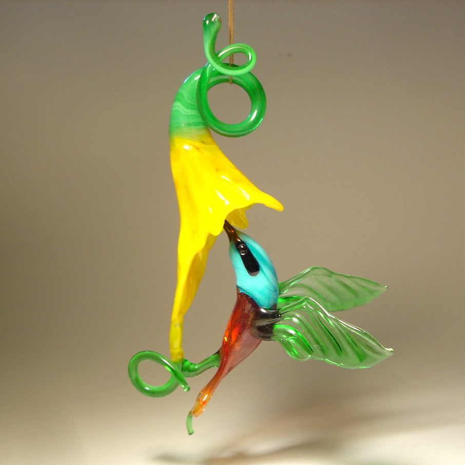 Suspended glass ornament featuring a blue head hummingbird in flight with a yellow trumpet flower.