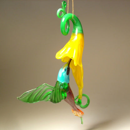 Glass hummingbird in green and red with a blue head under a yellow trumpet flower.