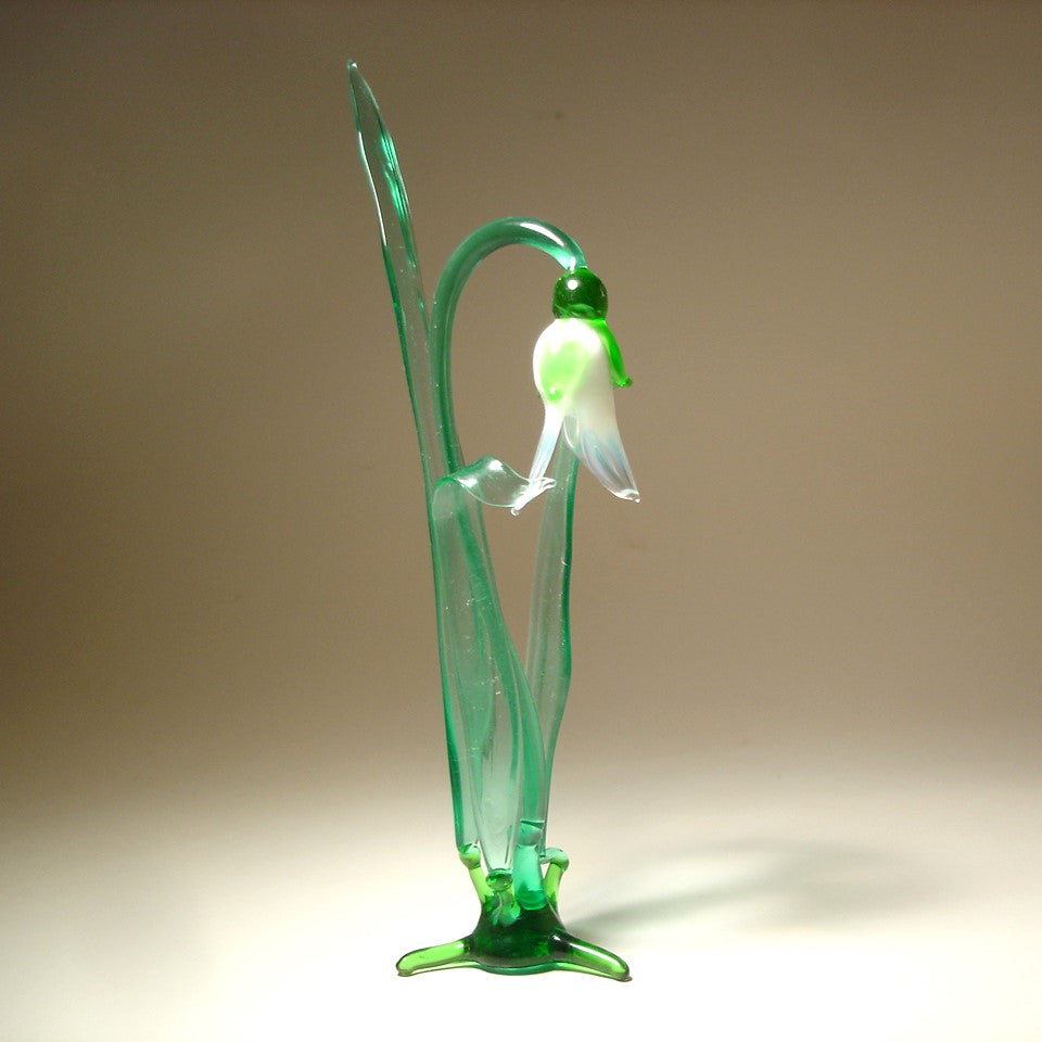 A front view of a delicate glass snowdrop figurine, showcasing its white petals and slender green stem.
