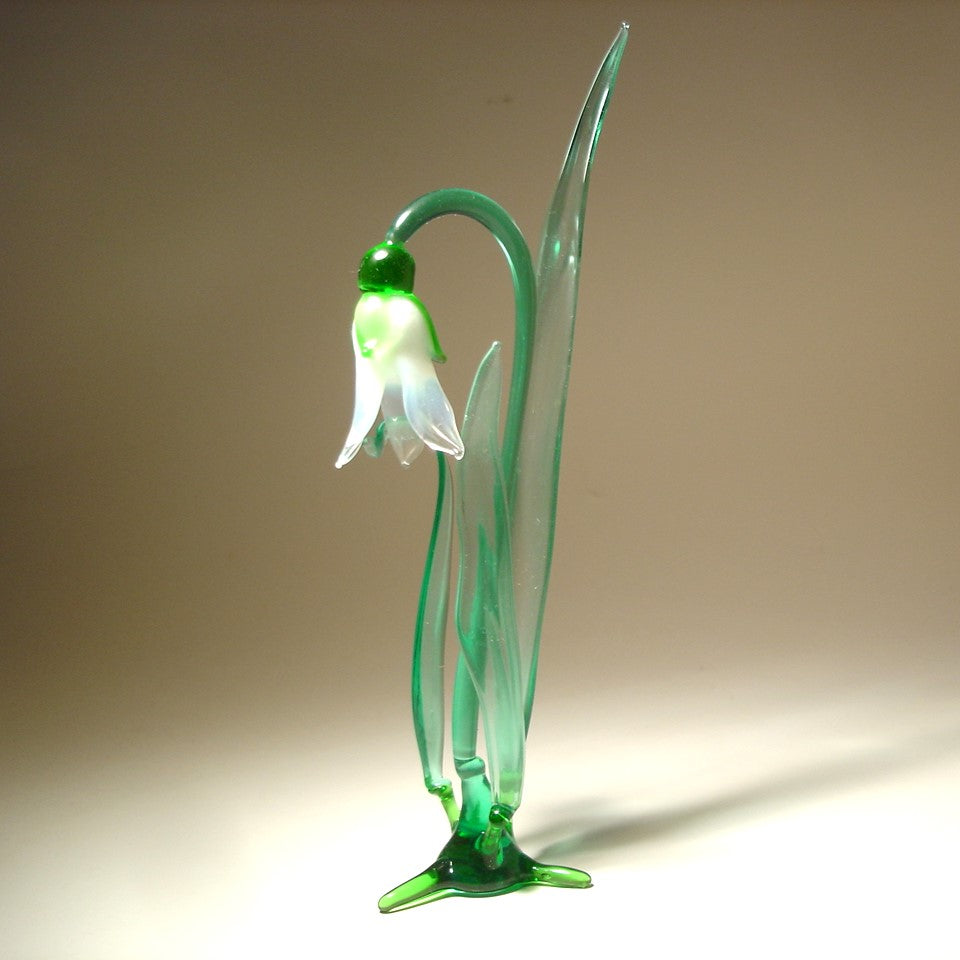 A side view of a glass snowdrop figurine, emphasizing the elegant curves of the petals and the smooth texture of the green stem.