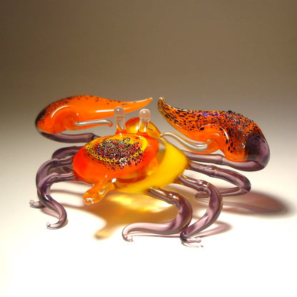 Glass sculpture of a crab with orange, purple, and yellow colors on a beige background.
