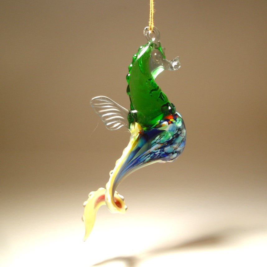 Side view of a glass green, blue and yellow seahorse ornament, focusing on the fine craftsmanship of its head, textured body, and hanging loop.