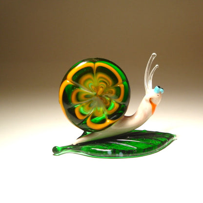 Aside view of a glass snail figurine resting on a leaf, emphasizing the spiral shape of its shell and the rich green color of the leaf.