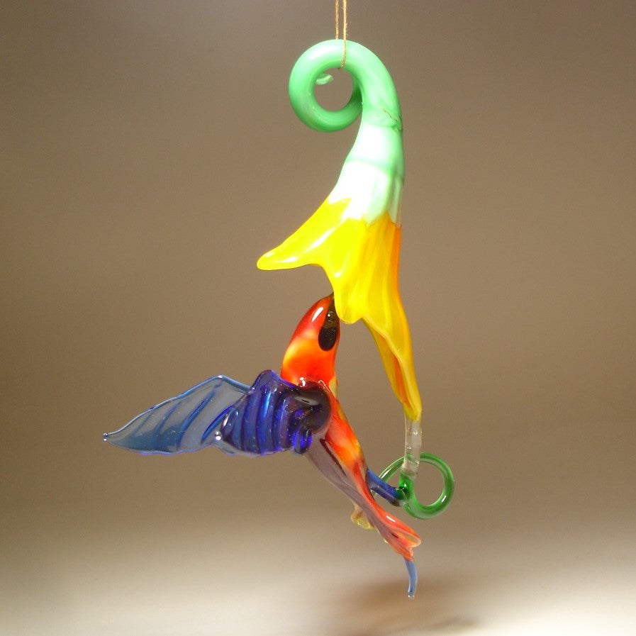 Suspended glass ornament featuring a red head hummingbird in flight with a yellow trumpet flower.