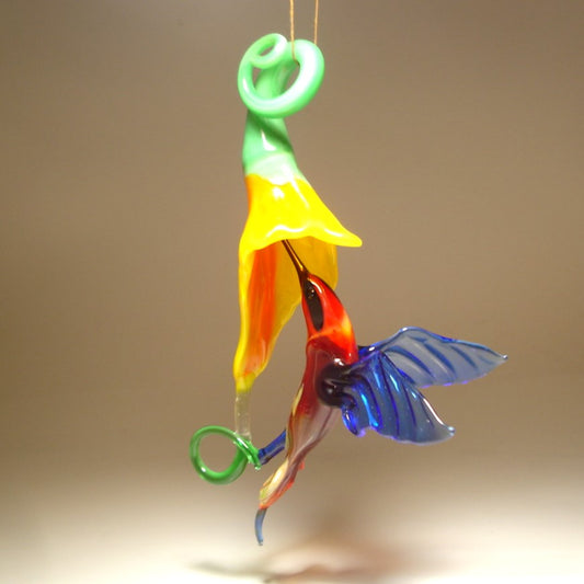 Glass hummingbird in green and red with a red head under a yellow trumpet flower.
