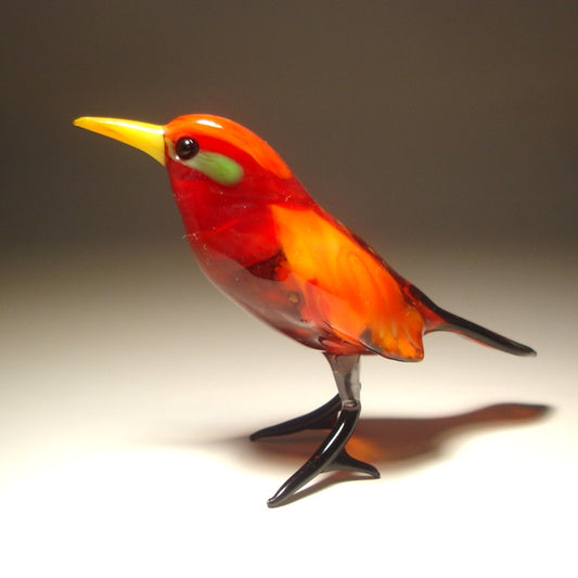 A handcrafted glass bird figurine with a vibrant red, orange, and yellow body, a bright yellow beak, and black legs, set against a dark gray gradient background.