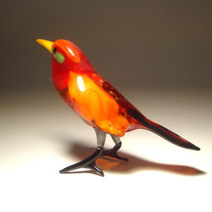 A hand-blown glass bird figurine with a vibrant red and orange body, a bright yellow beak, a small green eye patch, and black legs, set against a neutral gray background.