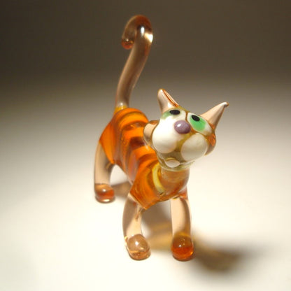 A handcrafted glass figurine of a cat in orange and white stripes with green and purple facial details, photographed on a neutral gradient background.