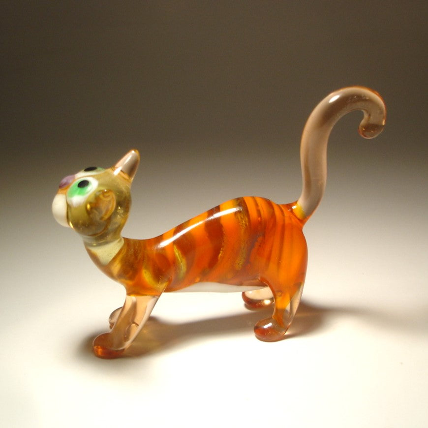 A hand-blown glass figurine of a walking cat in orange, amber, and cream tones with multicolored painted eyes, set against a dark-to-light gradient studio background.