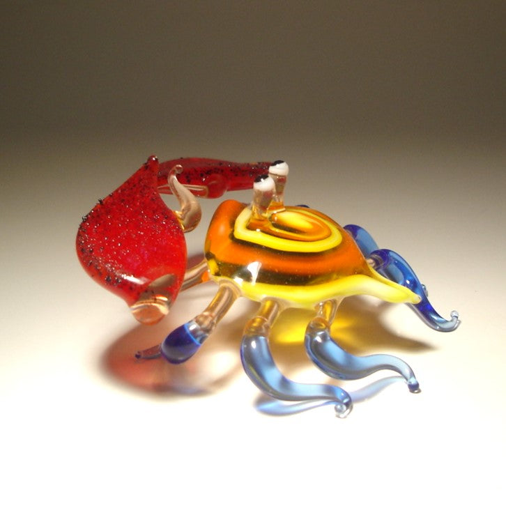 A hand-blown glass crab figurine with a red glittery claw, a yellow and orange body, and translucent blue legs.