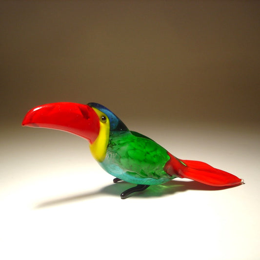 A side view of a glass toucan figurine, emphasizing the bold red beak and the smooth curves of its body.