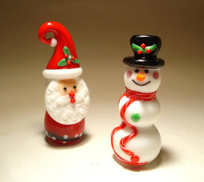 Side view of a glass snowman figurine, showcasing its carrot nose, coal eyes, and a colorful scarf around its neck. The Santa Clous figurine is standing further back.