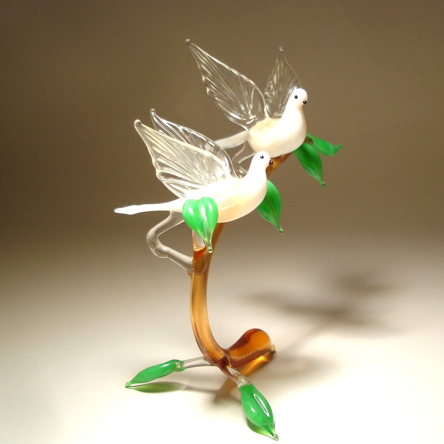 A decorative glass sculpture of two white doves perched on a curved brown stem with green leaves