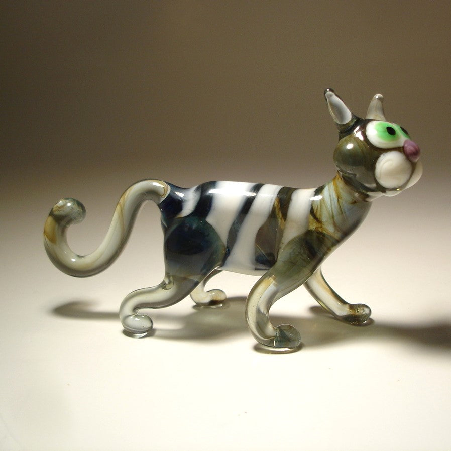 A glass figurine of a cat in a walking pose, featuring a gray and white striped body with green-tinted glass and bright green eyes.