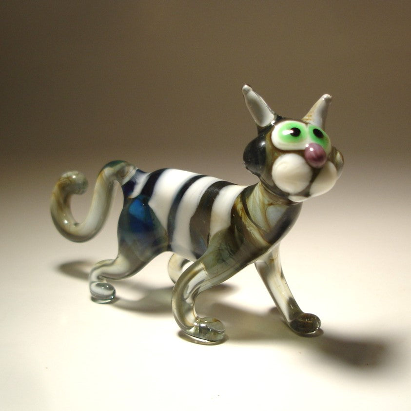 A glass figurine of a cat in a walking pose, featuring a gray and white striped body with green-tinted glass and bright green eyes.