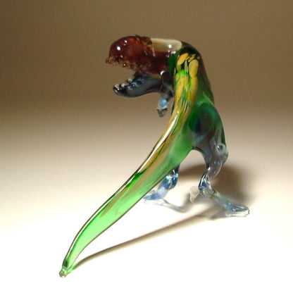 A hand-blown glass dinosaur figurine, likely a T-rex, featuring a translucent green body, blue legs, a dark blue back, and a reddish-brown head.