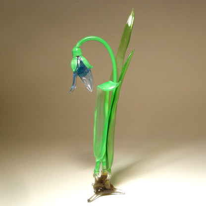 A hand-blown glass sculpture of a bell-shaped flower with a bright green stem and leaves, a blue-green bloom, and a clear glass base.