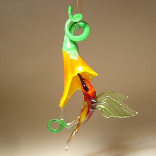 Glass hummingbird in green and red with a red head under a yellow trumpet flower.