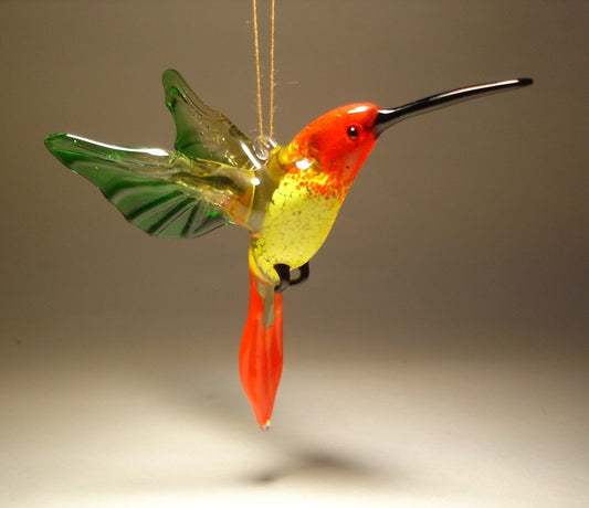 A vibrant red and yellow glass hummingbird ornament hanging from a delicate string, with green wings and a long pointed beak.