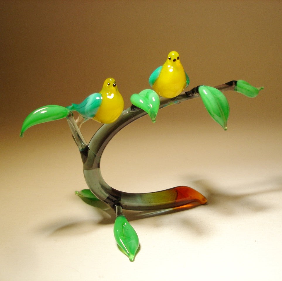 A glass figurine featuring two yellow with blue wings birds perched on a grey glass branch with leaves.