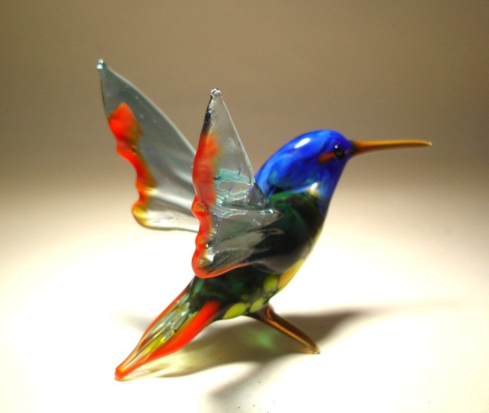 Close-up of a glass hummingbird figurine with a vibrant blue head, green body and light green wings trimmed in red.