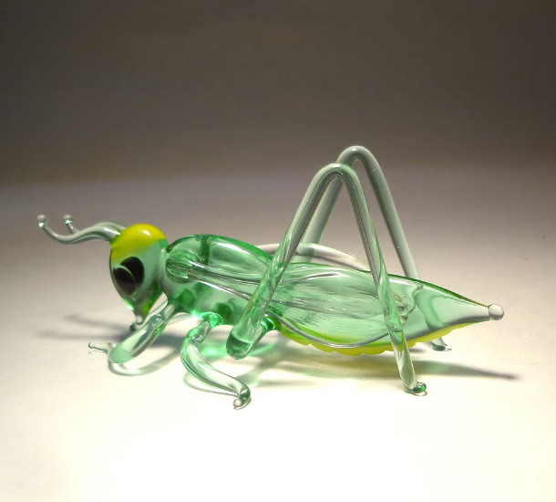 Left side view of a glass cricket grasshopper figurine, displaying its delicate legs, antennae, and light green body.