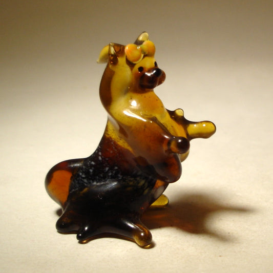 A charming glass Pekingese figurine in a sitting position, showcasing its fluffy coat and distinctive flat face, with an orange bow n its head.