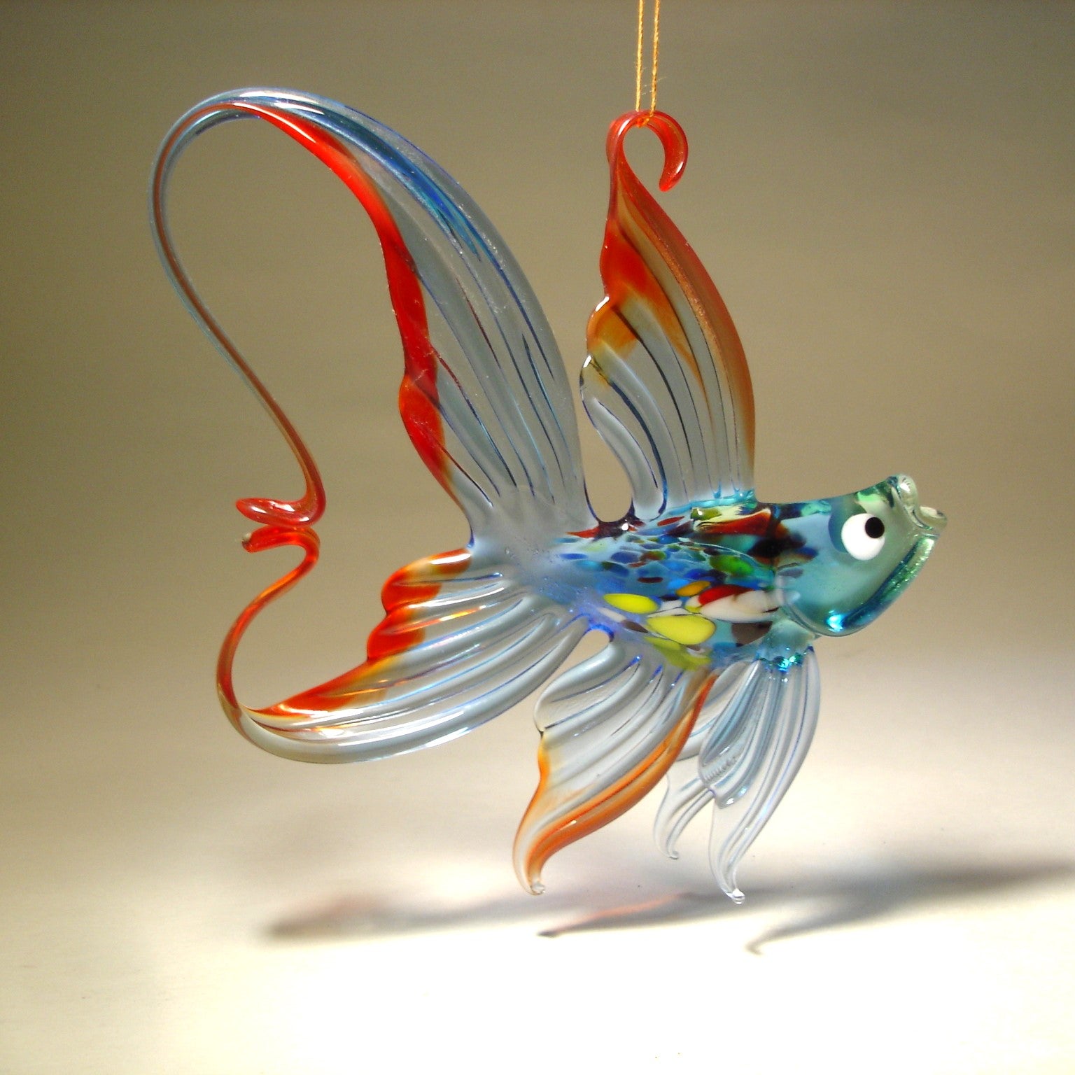 Side view of a suspended on a string handcrafted blue glass fish ornament with red trim around its fins