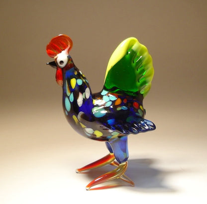 Side view of glass hen figurine with green tail trimmed in yellow and red feet.