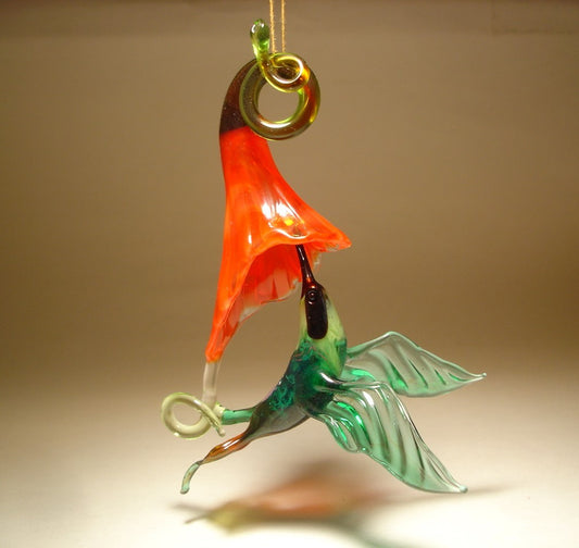 Glass hummingbird ornament in flight next to a bright red flower, showcasing the vibrant colors and intricate glasswork.