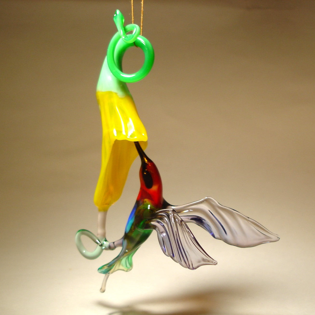 Suspended glass ornament featuring a red head hummingbird in flight with a yellow trumpet flower.