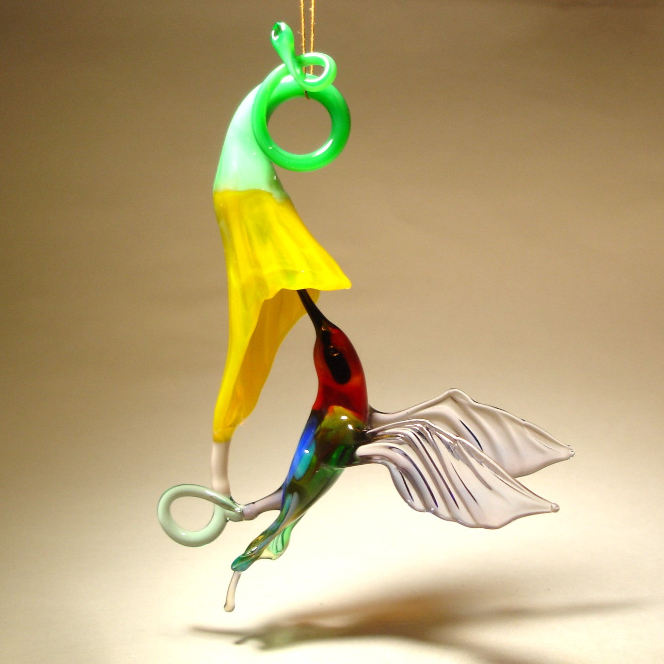 Glass hummingbird ornament hovering near a bright yellow flower, showing delicate details of the bird's wings and floral accents.